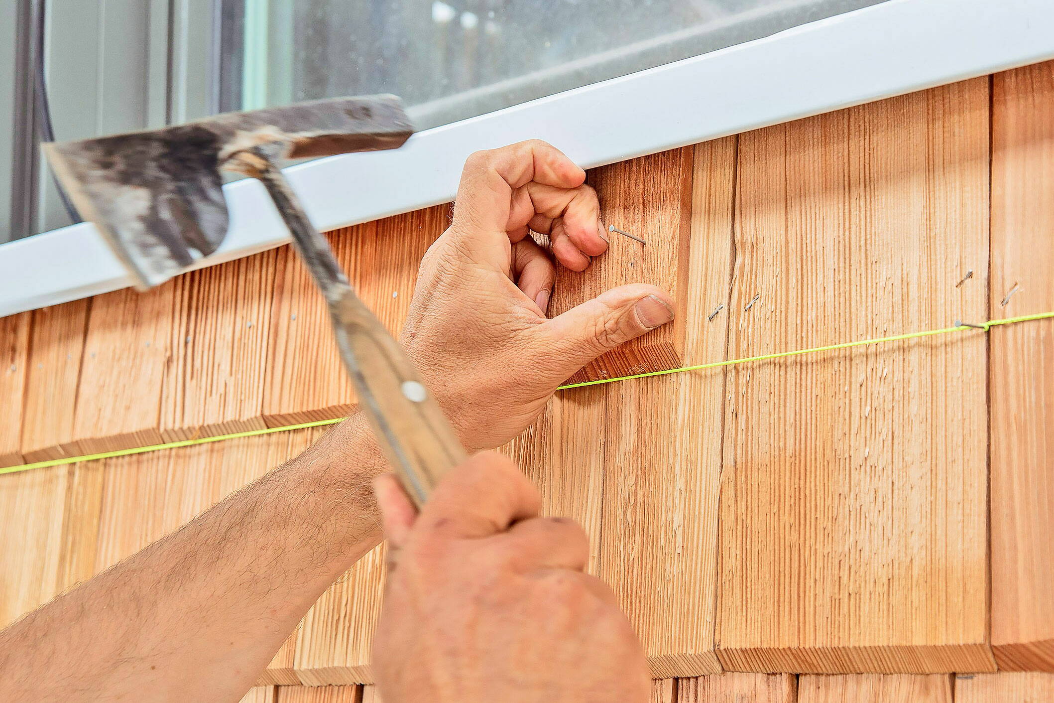 Ein Schindelspezialist ist dabei mit Hammer und Nagel eine neue Schindel an der Hauswand anzubringen.