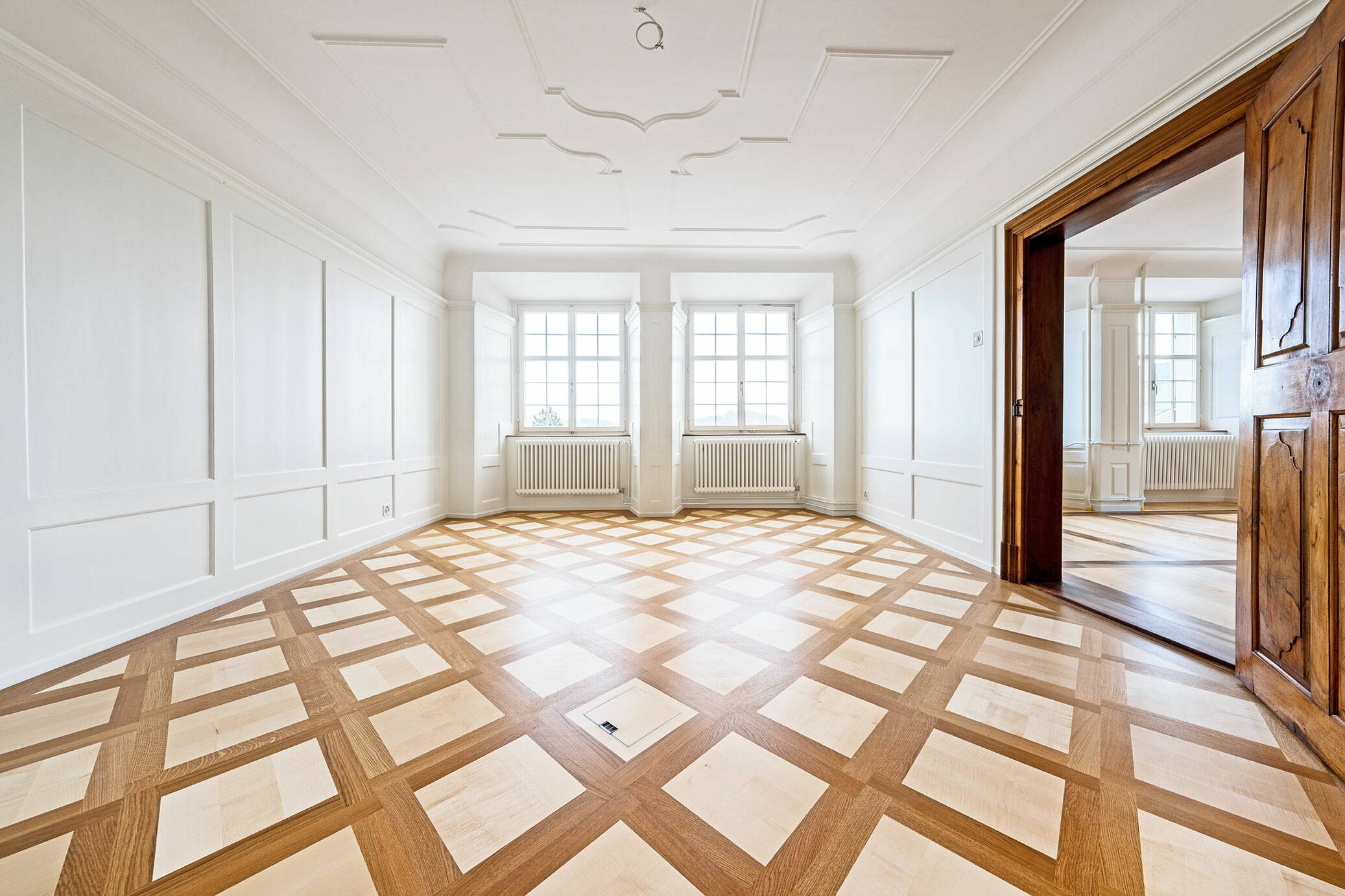 Sicht in einen lichtdurchfluteten Raum mit Holzparkett und Stuckaturen an der Decke.